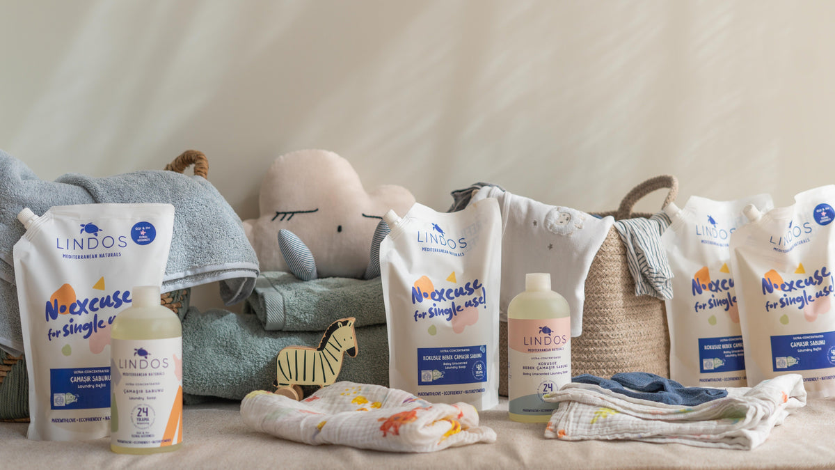 Lindos çamaşır sabunu şişeleri ve refill paketleri, yenidoğan kıyafetleri, havlu ve oyuncaklarla birlikte düzenlenmiş.